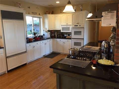Bright kitchen with white cabinets, double ovens, island stove, pendant lights, and wooden flooring.