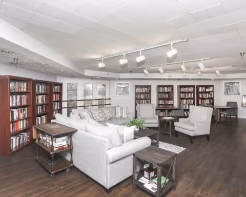 A spacious and well-lit common area with multiple bookshelves filled with books along the walls. The room features comfortable seating including a light gray sofa, armchairs, and small tables. The floor is dark wood, and the ceiling has track lighting with multiple spotlights. Large windows allow natural light to enter the room.