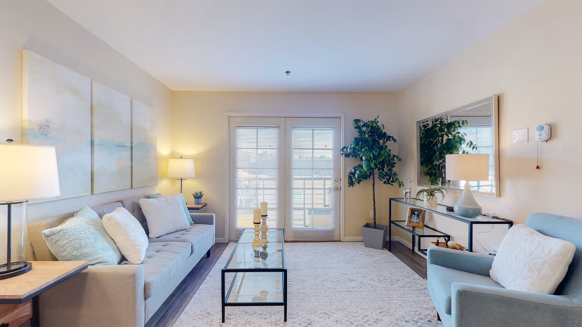 A bright and cozy living room with a light gray sofa adorned with white and light blue pillows, a glass coffee table with candles, a light blue armchair with a white pillow, a large mirror on the wall above a console table with a lamp and decorative items, a tall potted plant near French doors with blinds, and soft lighting from table lamps.