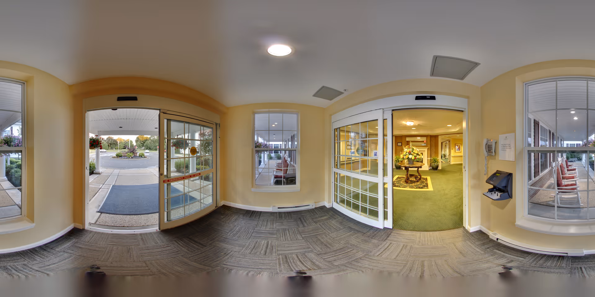 Entrance area of Brandywine Toms River by Monarch showing automatic sliding glass doors leading outside to a parking lot and inside to a lobby with green carpet, a round table with a flower arrangement, and seating visible through large windows.