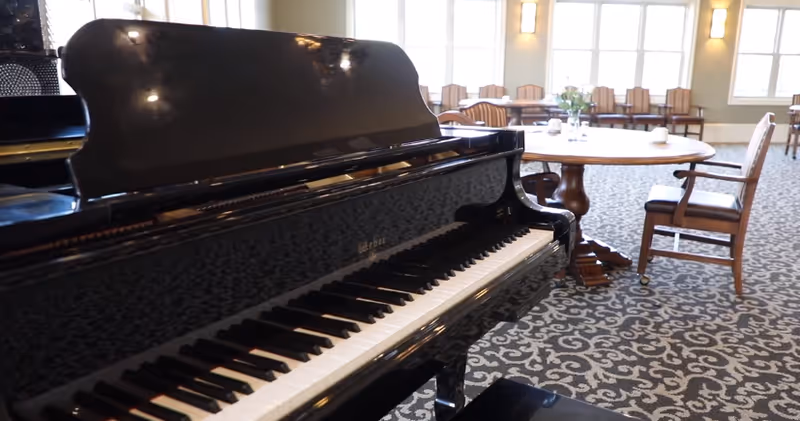 A black grand piano in the foreground of a dining/activity room with round tables, chairs, patterned carpet, and large windows.