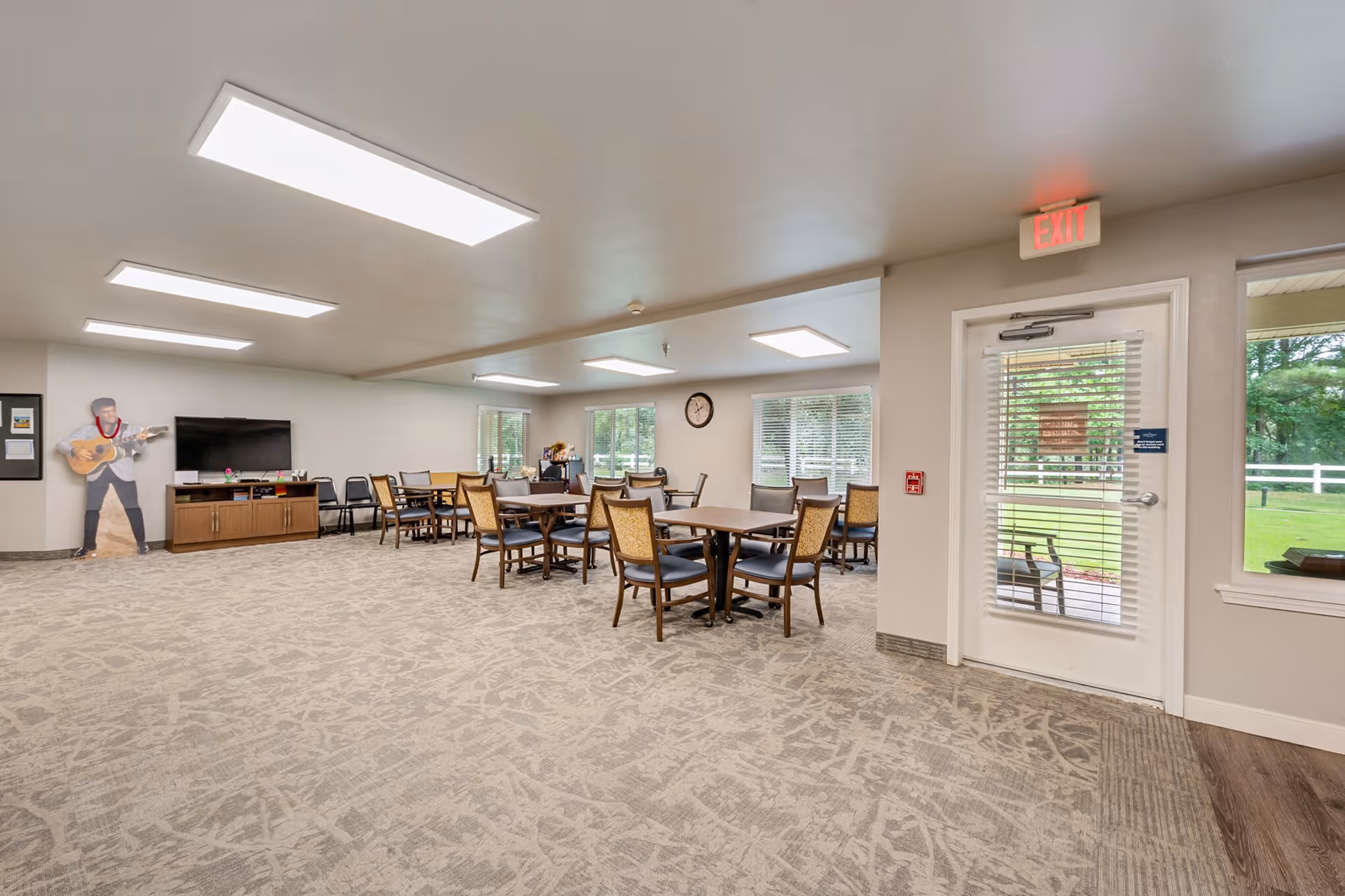A spacious common area with multiple tables and chairs arranged for group seating. The room has large windows letting in natural light, a television mounted on a wooden cabinet, and a cardboard cutout of a person playing guitar in the corner. The carpeted floor and neutral-colored walls create a welcoming atmosphere. An exit door with glass panels leads outside to a green outdoor space.