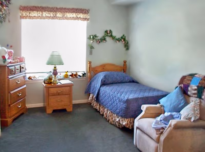 A cozy bedroom with a single wooden bed covered in a blue quilt, a wooden nightstand with a lamp and decorative items, a wooden dresser with stuffed animals on top, and a comfortable armchair with pillows and a blanket. A window with a valance lets in natural light, and there is a floral garland decoration on the wall above the bed.