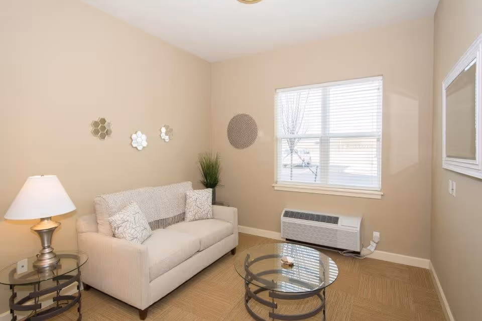 A small living room with beige walls and carpet. There is a light beige sofa with two patterned pillows, a glass-top round coffee table with a decorative item, and a glass side table with a lamp. A window with white blinds lets in natural light, and a wall-mounted heater is below the window. Decorative wall mirrors and a plant add accents to the room.