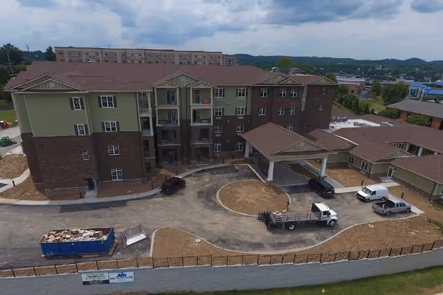 Aerial view of a multi-story senior living facility building with green and brown exterior walls, a covered entrance driveway, several parked vehicles, and surrounding landscaping under construction.