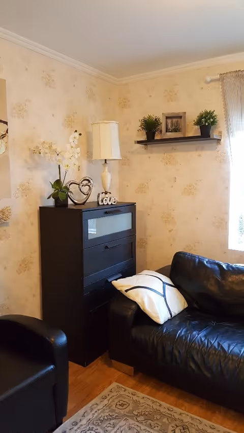 A cozy living room corner with a black leather sofa and a matching armchair. A black cabinet with decorative items including a white lamp, a heart-shaped ornament, a 'Love' sign, and a potted orchid sits against a floral wallpapered wall. Above the cabinet, a floating shelf holds two small potted plants and a framed picture. A window with striped curtains allows natural light into the room.