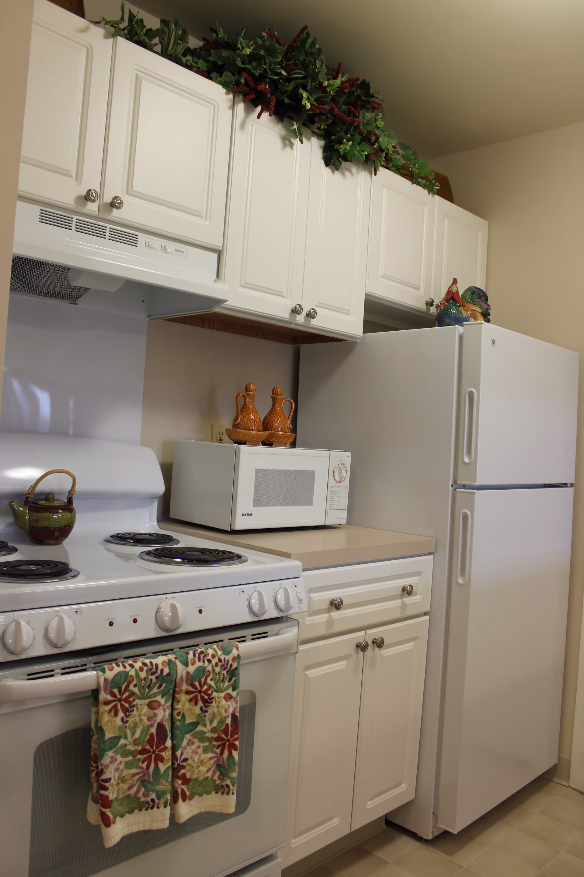 A kitchen area with white cabinets, a white electric stove with floral patterned towels hanging on the handle, a microwave on the countertop, and a white refrigerator. Decorative greenery is placed above the cabinets, and ceramic pitchers and a rooster figurine are on the countertop and refrigerator.