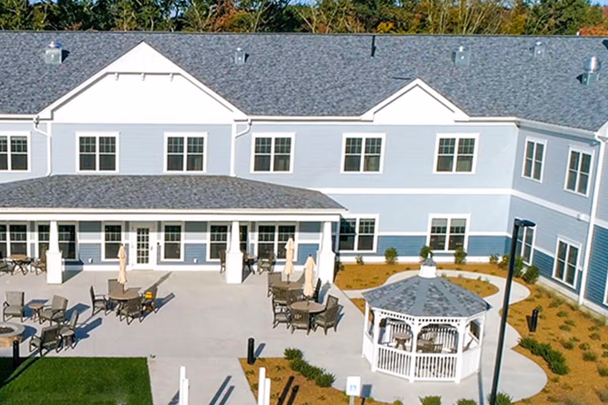 Outdoor patio area of a senior living facility with multiple tables and chairs, umbrellas, and a white gazebo. The building in the background is two stories with blue and white siding and multiple windows. There is a concrete walkway and landscaped areas with small bushes and mulch.