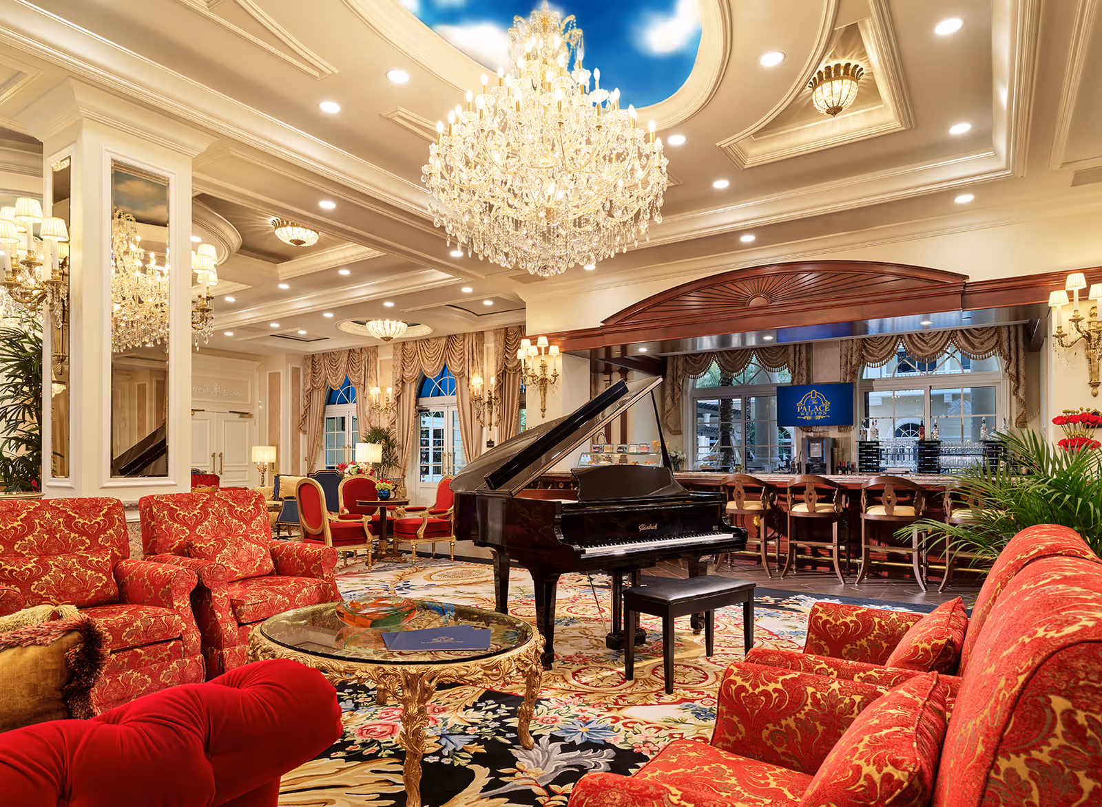 Opulent communal lounge with red upholstered seating, a grand piano under crystal chandeliers, and a bar in the background.