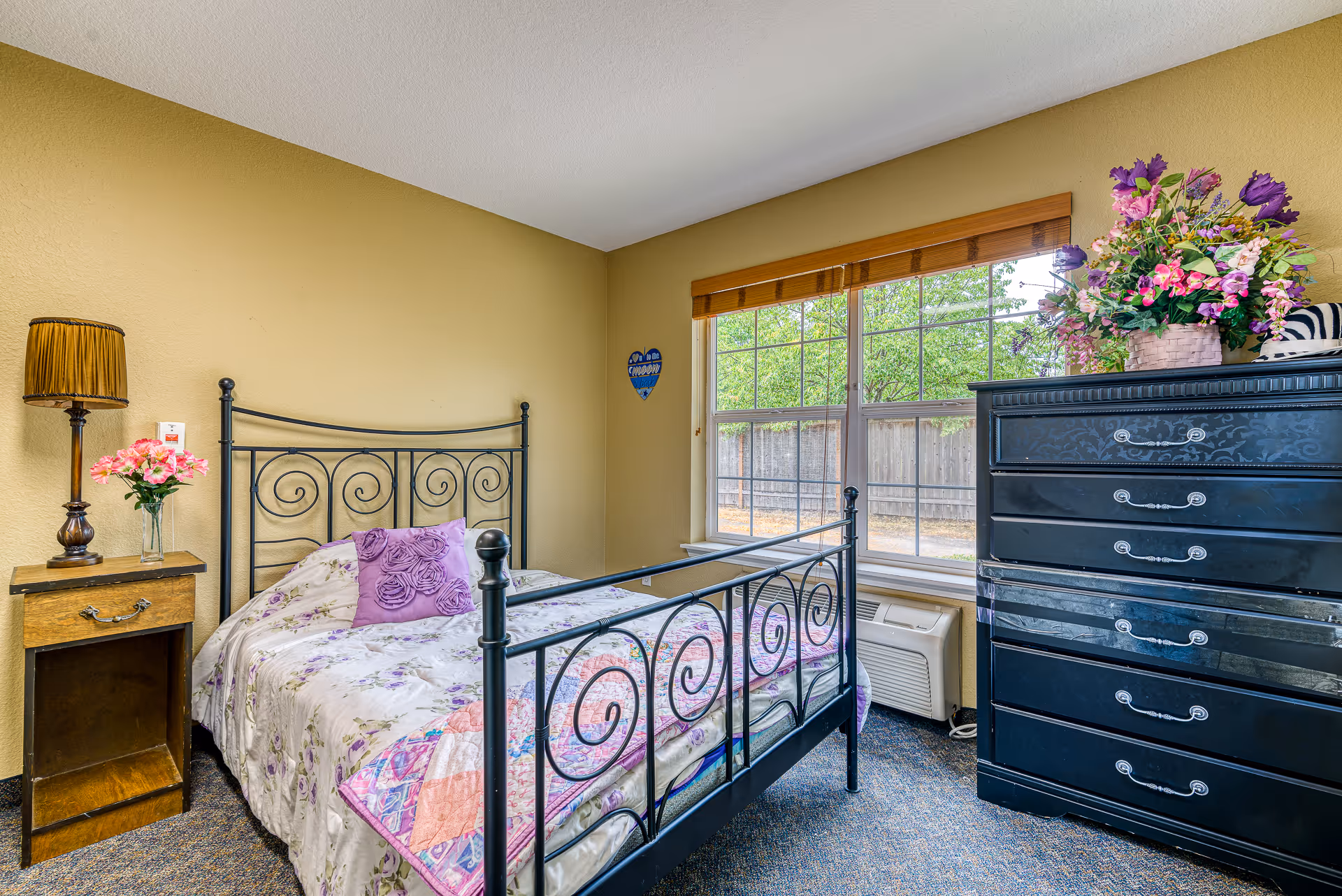 A cozy bedroom with a black metal bed frame featuring spiral designs, a floral bedspread with a purple decorative pillow, a wooden nightstand with a vintage lamp and a vase of pink flowers, a large window with wooden blinds showing a fenced outdoor area, and a black dresser topped with a basket of colorful artificial flowers.