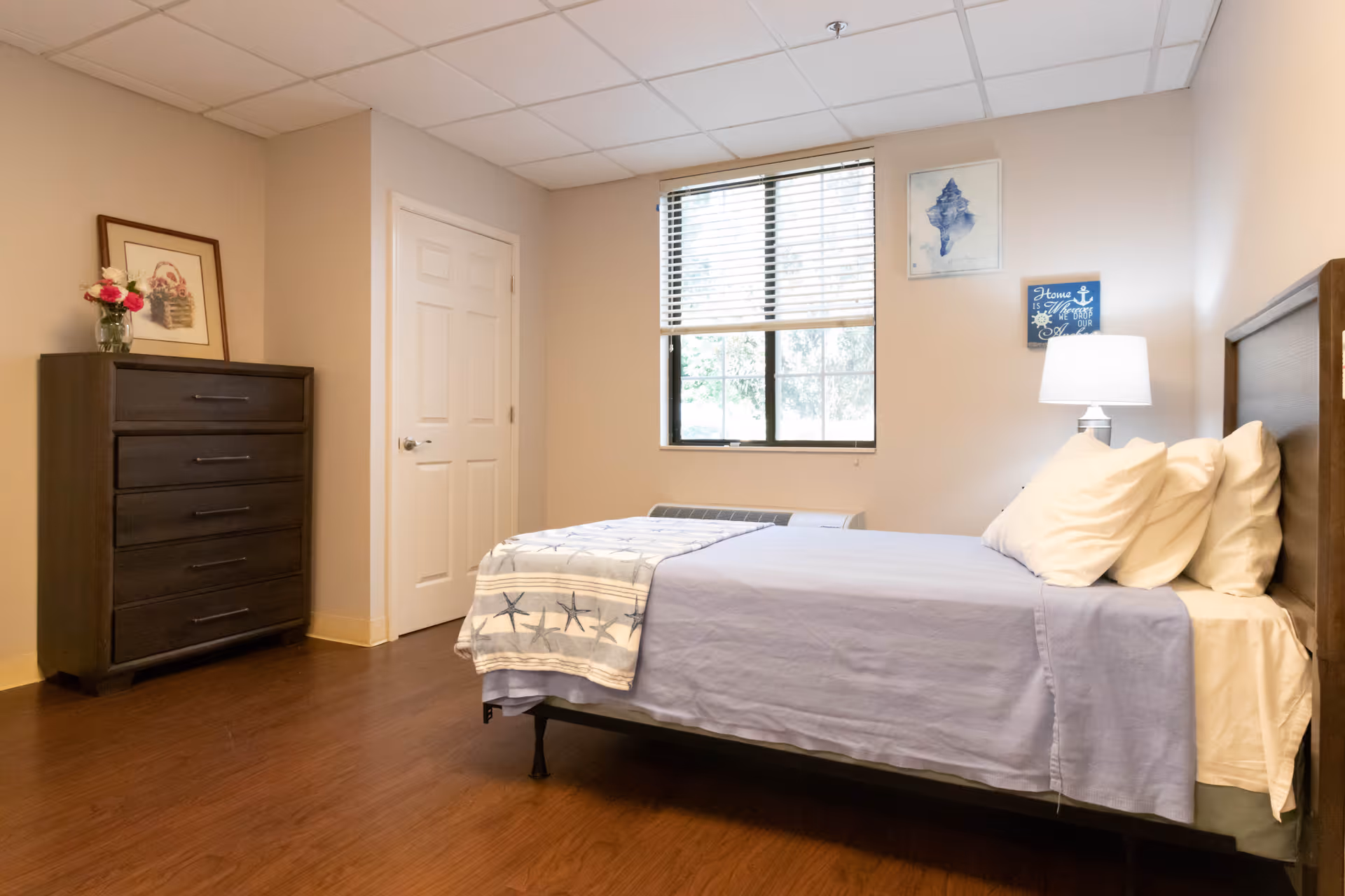 A neatly made bed with light blue bedding and multiple pillows in a senior living facility bedroom. The room has wooden flooring, a window with blinds, a dark wooden dresser with a framed picture and a vase of flowers on top, and a bedside lamp on a nightstand. The walls are light-colored with two pieces of wall art.