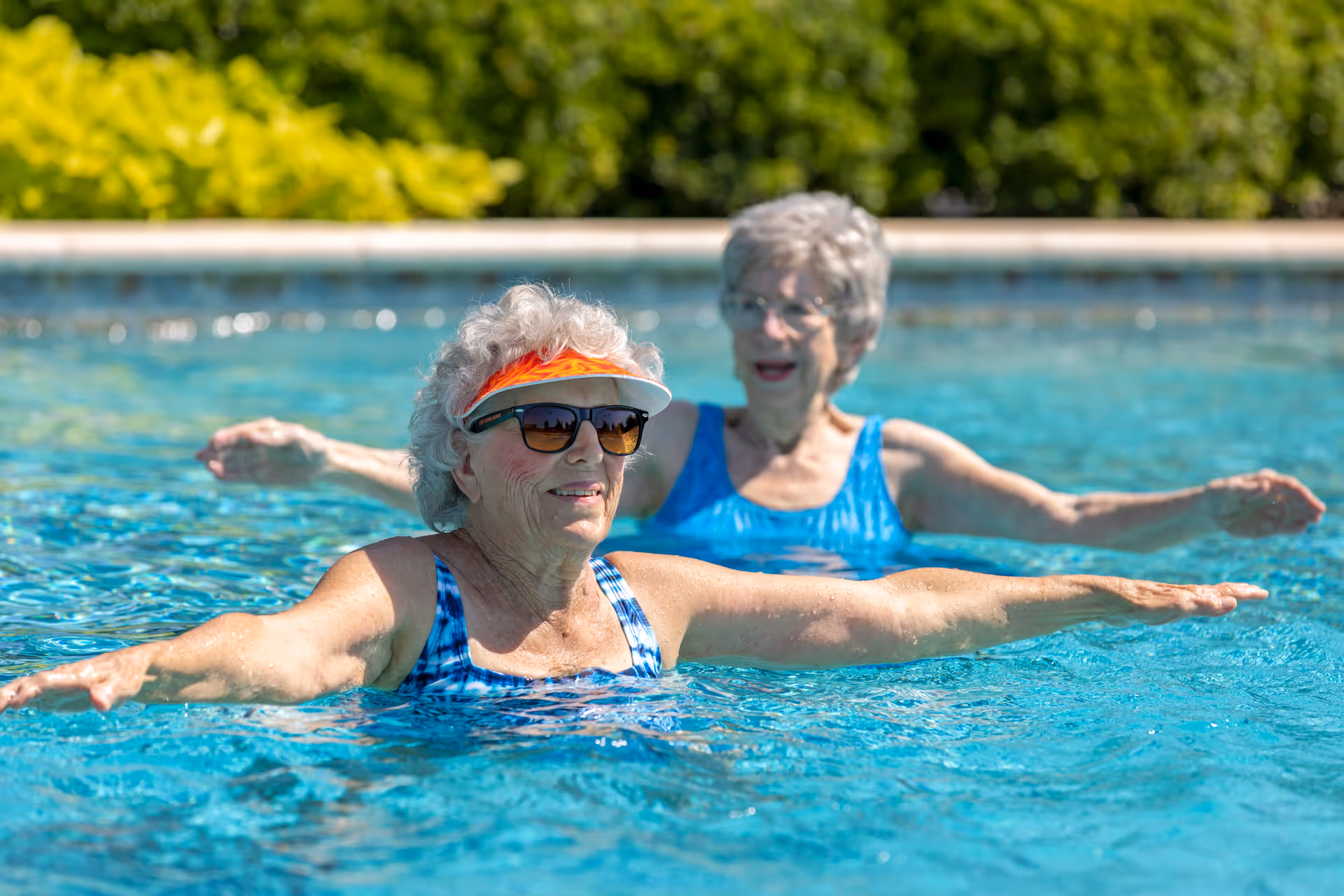 Two elderly women in blue swimsuits exercising in an outdoor swimming pool with greenery in the background. One woman is wearing sunglasses and an orange visor.