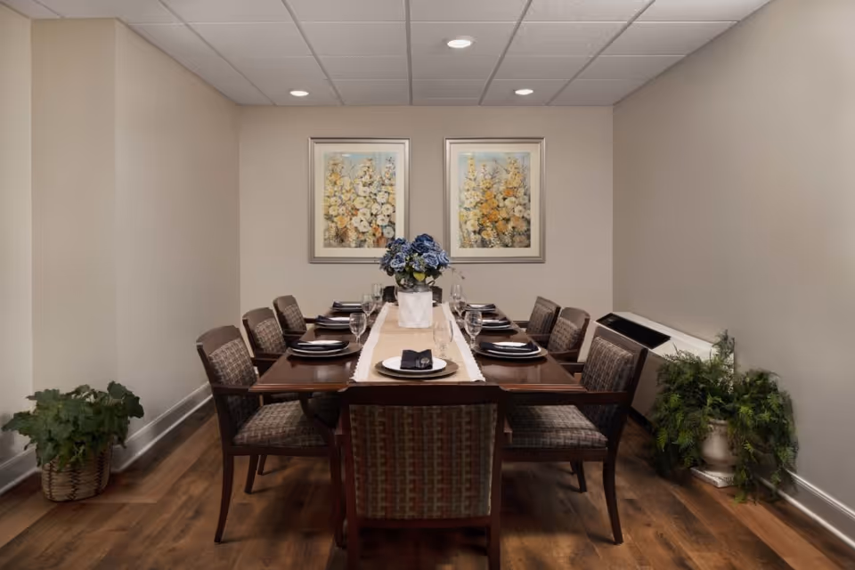 A dining room with a rectangular wooden table set for eight people. Each place setting includes a plate, napkin, and glass. A white table runner with a vase of blue flowers is centered on the table. Two framed floral paintings hang on the beige wall behind the table. There are two potted plants on the floor, one on the left and one on the right side of the room. The floor is wooden, and the ceiling has recessed lighting.