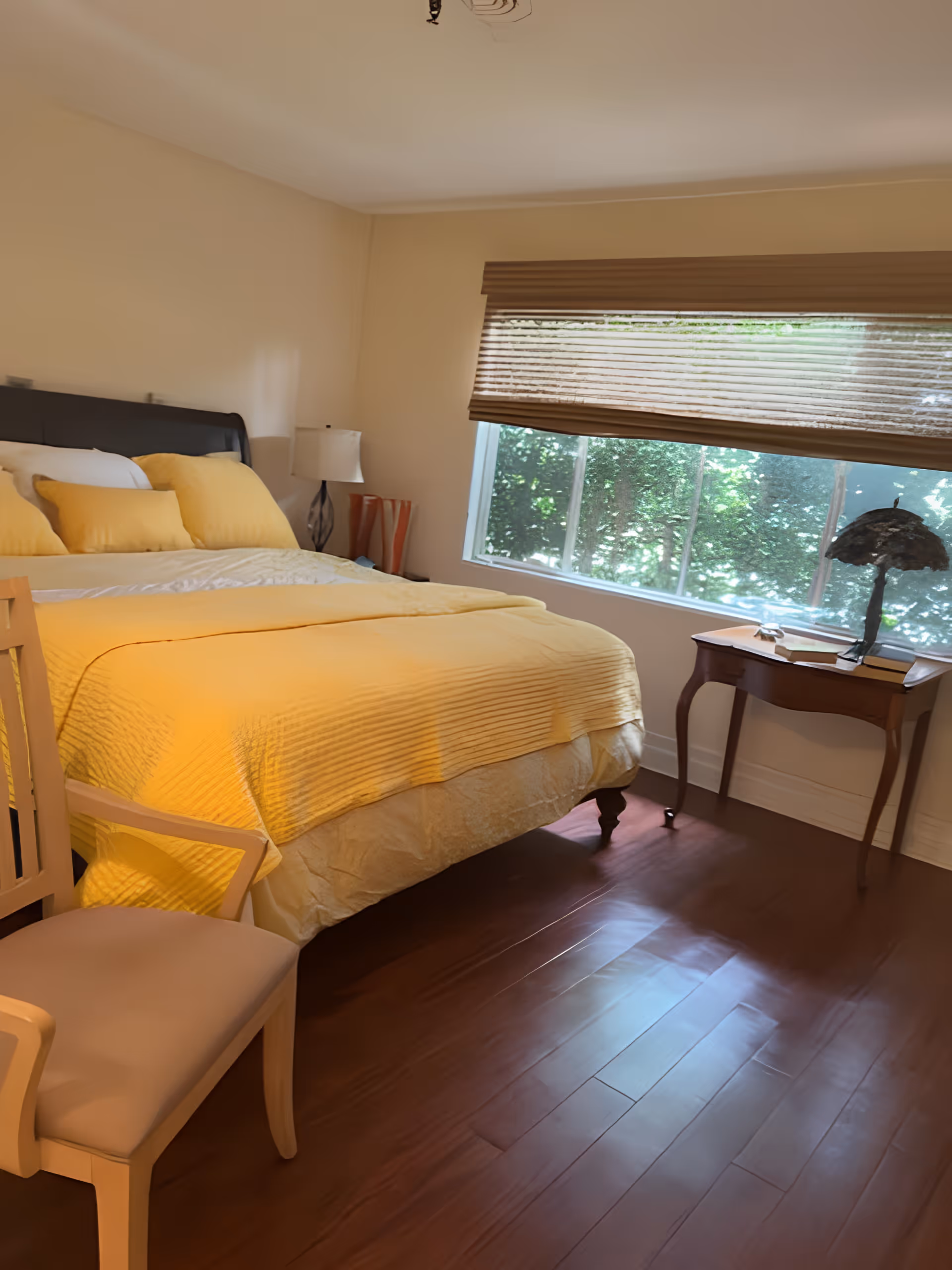 A bedroom with a large bed covered in yellow bedding and multiple pillows. There is a wooden chair with a cushioned seat in the foreground, a small wooden side table with a decorative lamp and books near a window with partially closed blinds, and hardwood flooring.