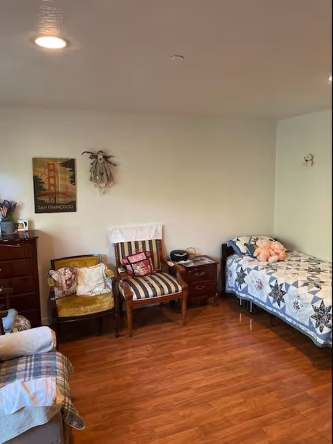 A cozy room with wooden flooring featuring a single bed with a quilt and stuffed animals, two vintage chairs with cushions, a small wooden nightstand with a telephone, and a dresser. The walls are plain with minimal decorations including a San Francisco poster and a hanging doll.