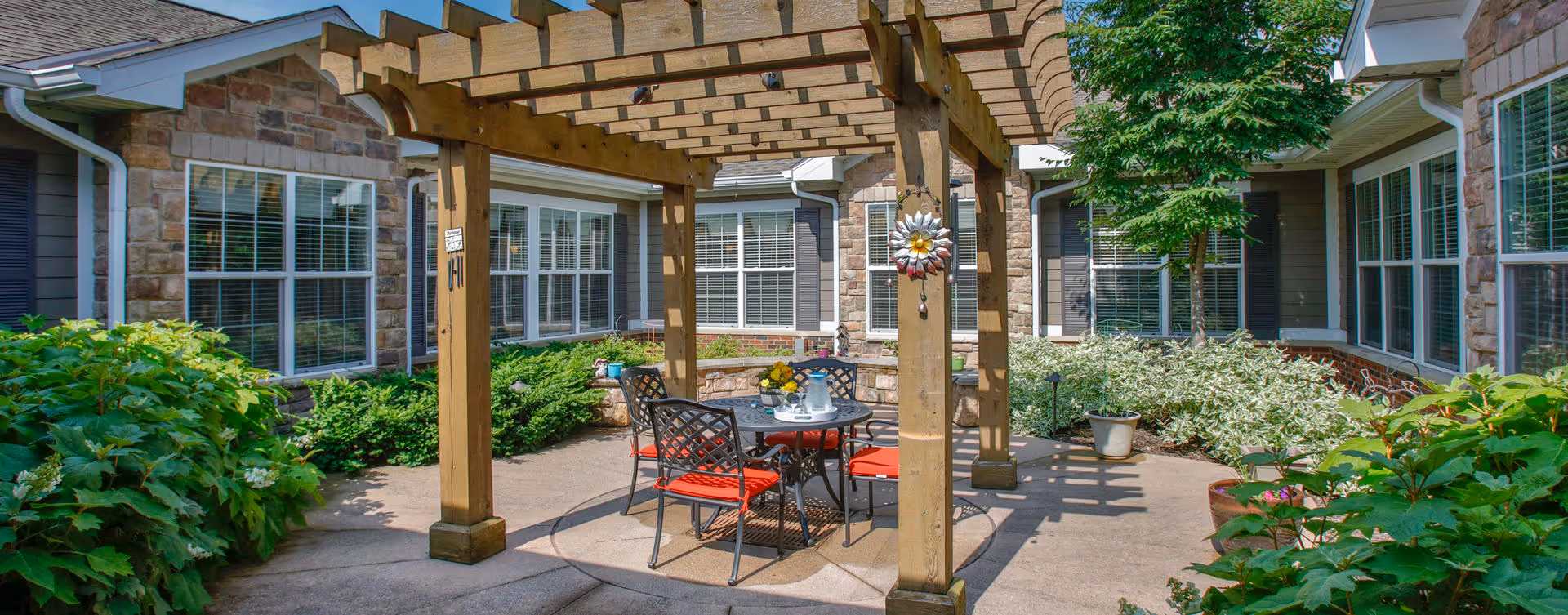 Outdoor patio area at Bickford of Greenwood featuring a wooden pergola with a round table and four chairs with red cushions underneath. The patio is surrounded by green shrubs, potted plants, and the building's stone and siding exterior with multiple windows.