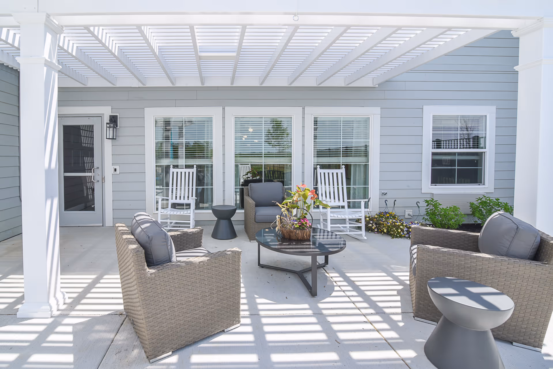 Outdoor patio area with a white pergola overhead casting shadows on the concrete floor. The space includes two cushioned wicker armchairs, two white rocking chairs, a small black side table, a round black coffee table with a flower arrangement, and some greenery along the building wall with light gray siding and white trim around the windows and door.