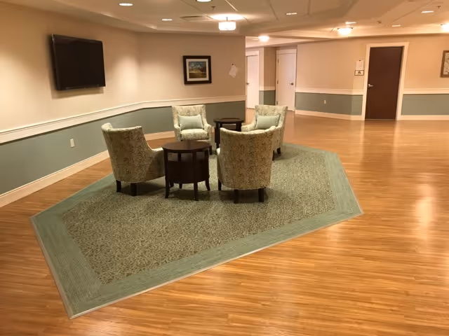 A seating area in a hallway with four upholstered armchairs arranged around two small round wooden tables on a patterned area rug. The hallway has wood flooring, beige and green walls with white trim, a wall-mounted flat screen TV, framed artwork, and several closed doors.