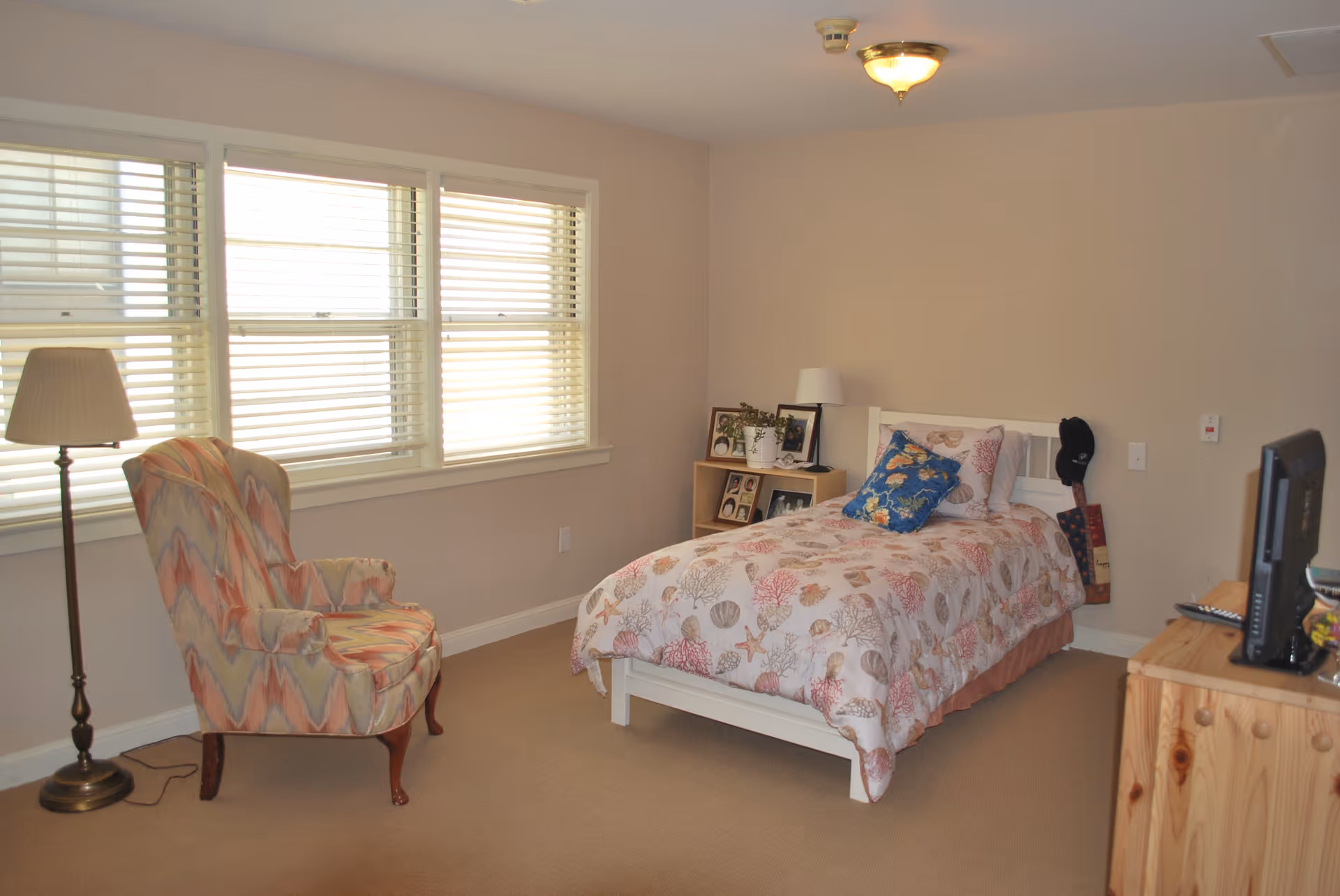 Bright bedroom with a twin bed, patterned armchair, floor lamp, dresser with a TV, and large windows with blinds.