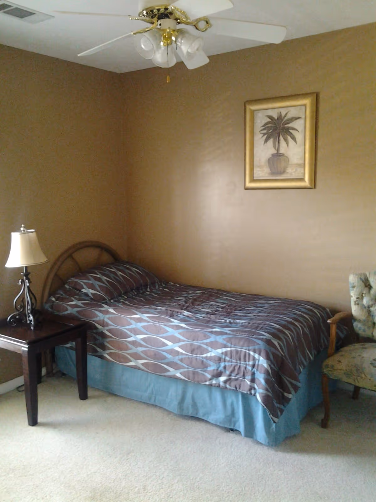 A simple bedroom with a bed covered in patterned bedding, a nightstand with a lamp, a ceiling fan, framed wall art, and an armchair.
