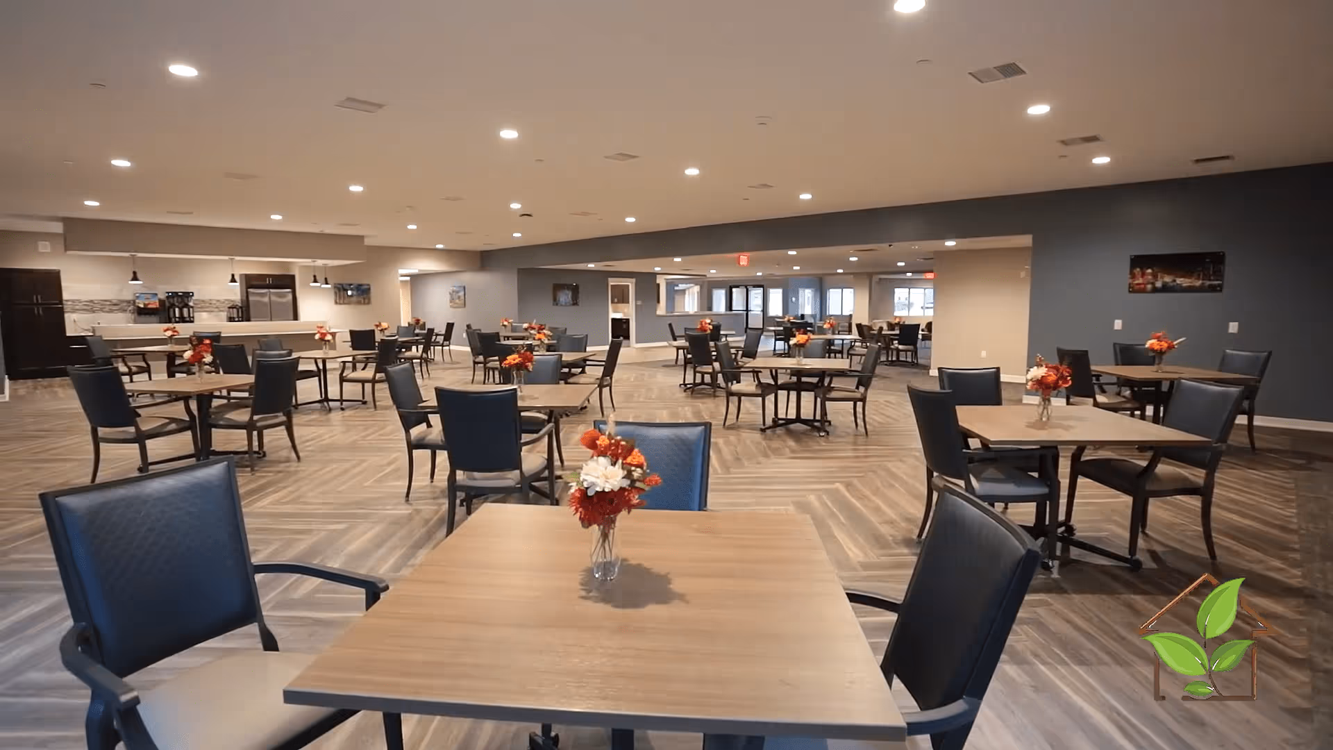 A spacious dining room with multiple square wooden tables, each adorned with a small vase of red and white flowers. The room has modern lighting with recessed ceiling lights, a patterned floor, and a kitchen area with refrigerators and coffee machines in the background.