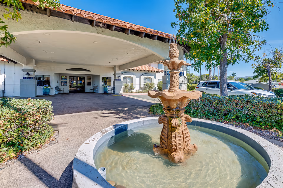 Outdoor view of the entrance to Solstice Senior Living at El Cajon featuring a covered driveway, a decorative water fountain in the foreground, surrounding greenery, and parked cars under a clear blue sky.
