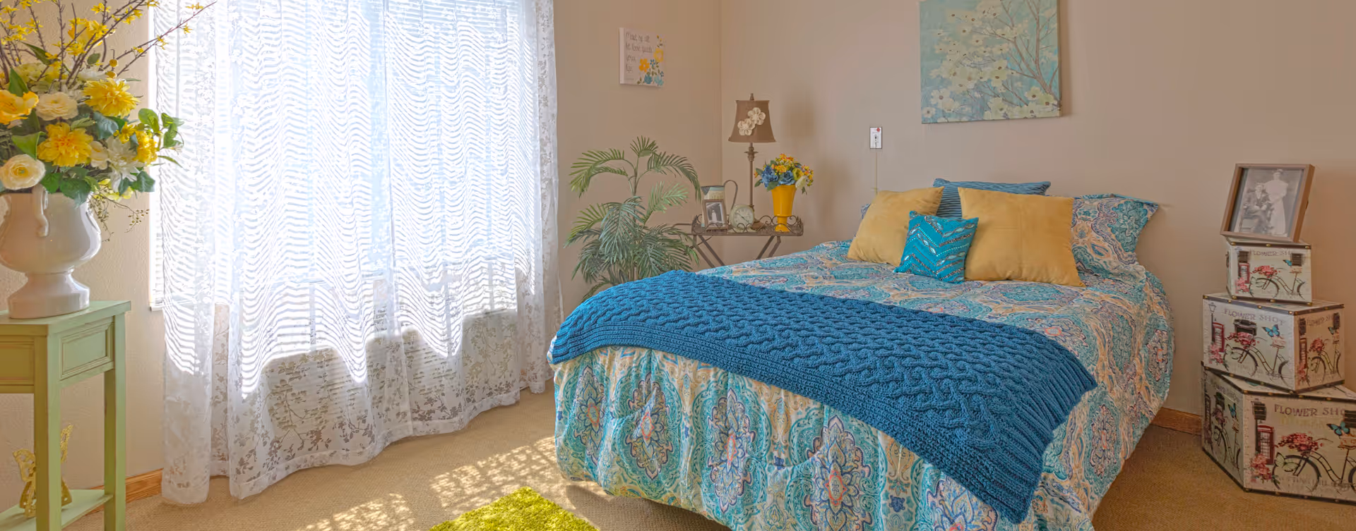 Senior living bedroom with patterned quilt, teal throw, plants, and lace-curtained window.