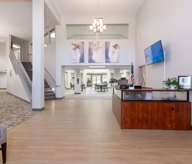 Spacious and well-lit interior of a senior living facility with a wooden reception desk on the right, a mounted flat-screen TV above it, and an American flag beside the desk. The area features light wood flooring, white walls, and a staircase with carpeted steps on the left. In the background, there are tables and chairs arranged in a common area with large windows letting in natural light. Three abstract paintings hang on the wall above the entrance to the common area.