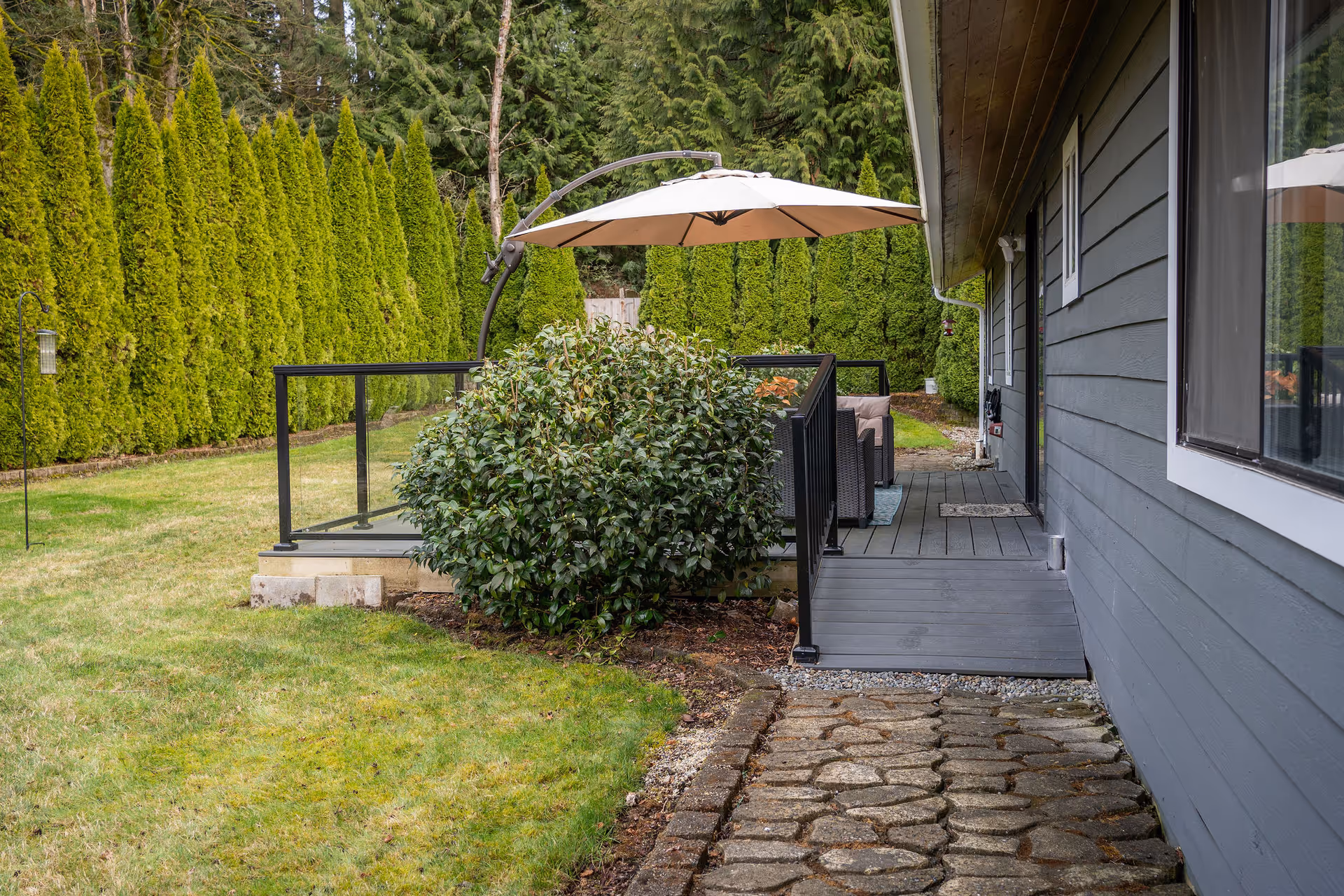 Outdoor patio area next to a gray building with a stone pathway leading to a deck. The deck has black railing with glass panels, a large beige umbrella, and outdoor seating. The area is surrounded by green grass, a large bush, and tall evergreen trees in the background.