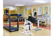 A physical therapy or rehabilitation room with exercise equipment including a treadmill, stationary bike, and other fitness machines. The room has large windows with blinds, yellow walls, and a clean, organized appearance.