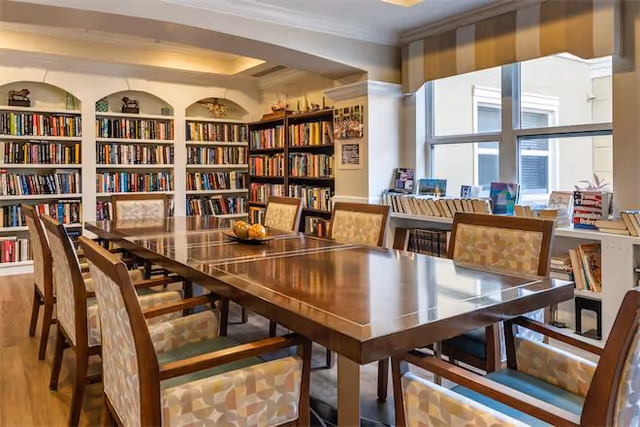 A well-lit room with a large wooden table surrounded by cushioned chairs with patterned upholstery. The walls are lined with bookshelves filled with books, and there is a large window letting in natural light. The room appears to be a library or reading area within a facility.