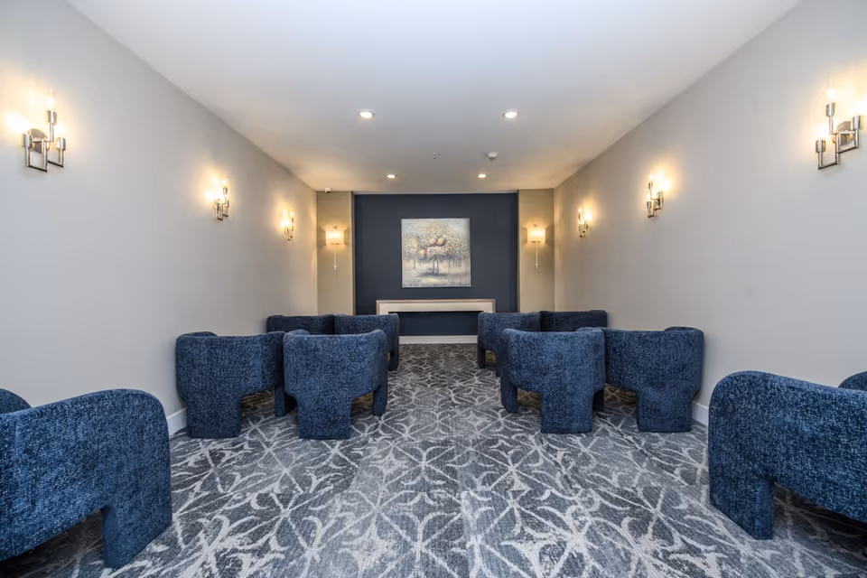 A small meeting or lounge room with blue upholstered chairs arranged in rows facing a wall with a painting of a tree. The room has light gray walls, patterned carpet, and wall-mounted light fixtures providing warm lighting.