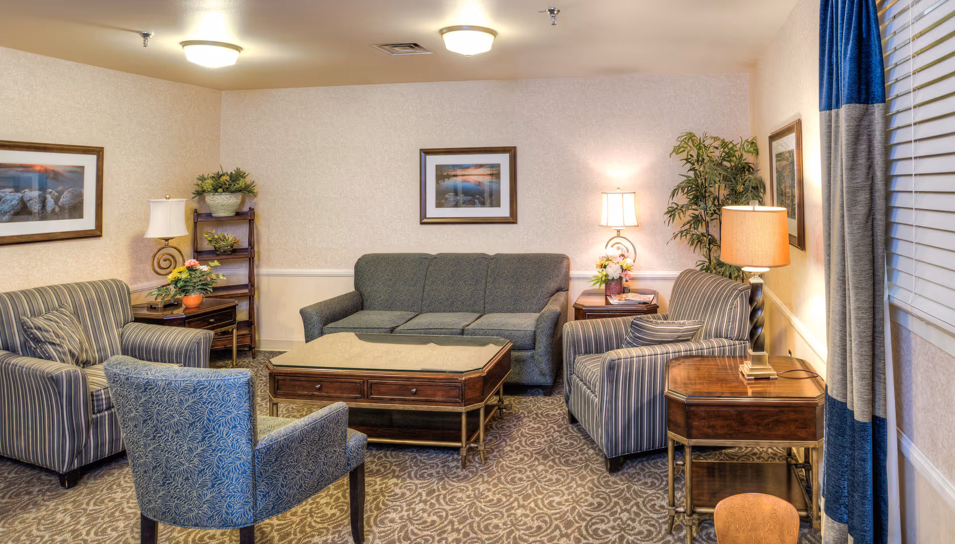 A cozy living room area with a gray sofa, two striped armchairs, and a blue patterned chair arranged around a wooden coffee table. The room has beige walls with framed landscape pictures, two table lamps on side tables, a floor lamp, a small wooden shelf with plants, a potted plant in the corner, and blue and gray curtains covering a window with blinds.