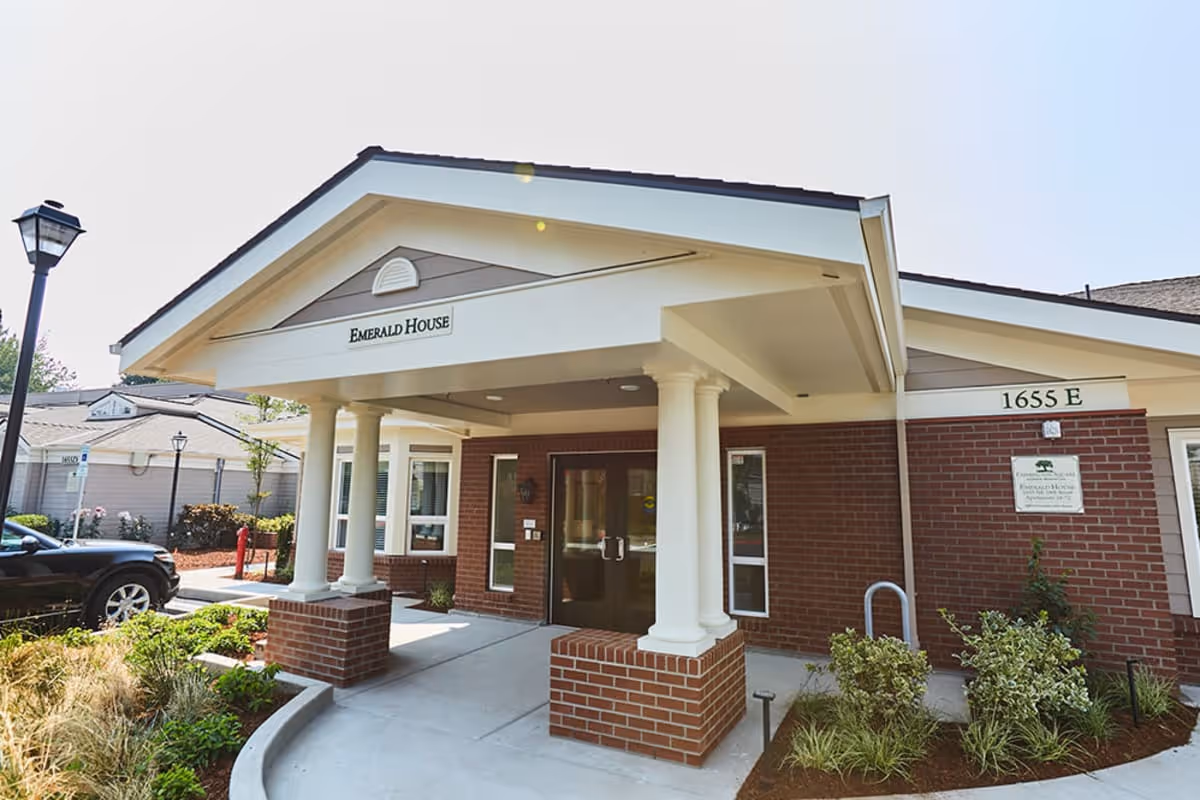 Front exterior view of a senior living facility entrance named Emerald House with brick walls, white columns, and a covered porch. There are plants and shrubs along the walkway and a black car parked nearby.