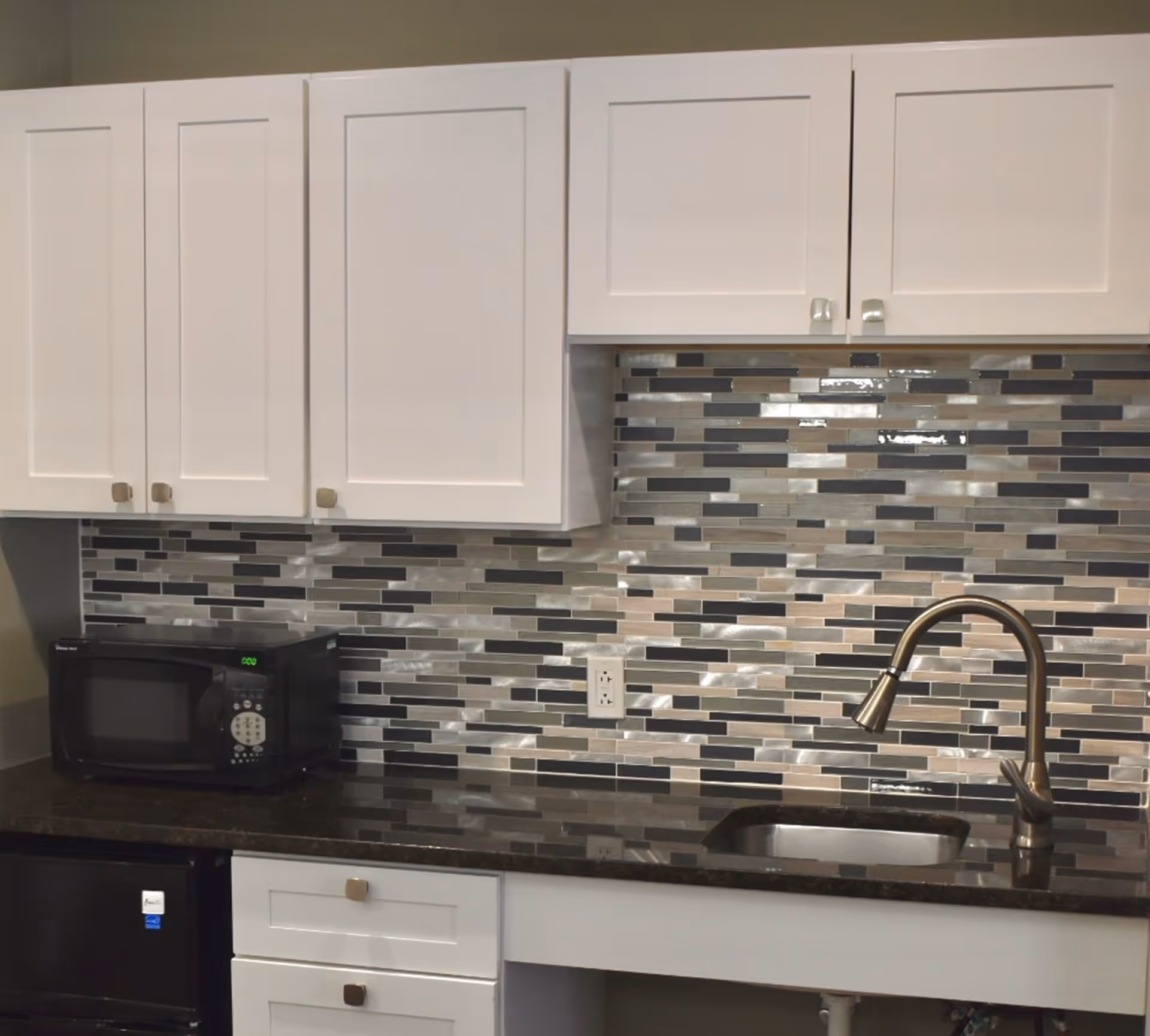 Compact kitchen counter with white cabinets, a multicolored glass tile backsplash, a microwave, sink, and a brushed metal faucet.