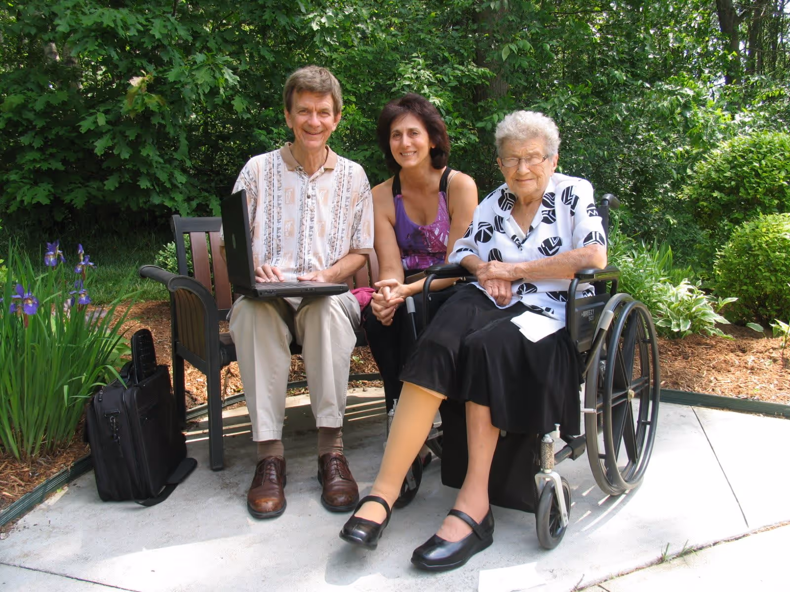 An elderly woman in a wheelchair, a middle-aged woman, and an elderly man sitting on a bench outdoors surrounded by greenery. The elderly man is using a laptop, and there is a black bag placed on the ground next to the bench.