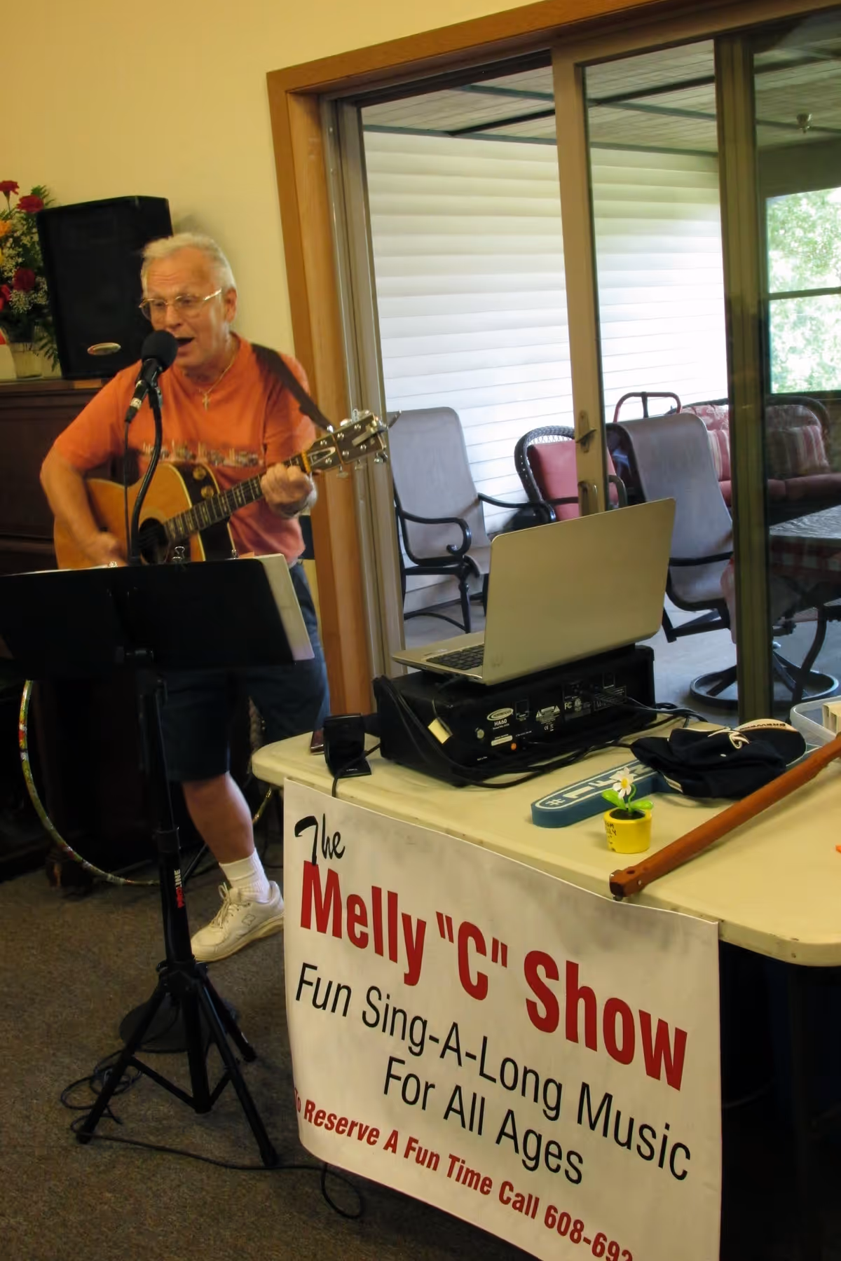 An elderly man wearing an orange shirt and shorts is playing an acoustic guitar and singing into a microphone in an indoor setting. In front of him is a table with a laptop, audio equipment, and a sign that reads 'The Melly "C" Show Fun Sing-A-Long Music For All Ages'. Behind him is a sliding glass door leading to a patio with chairs.