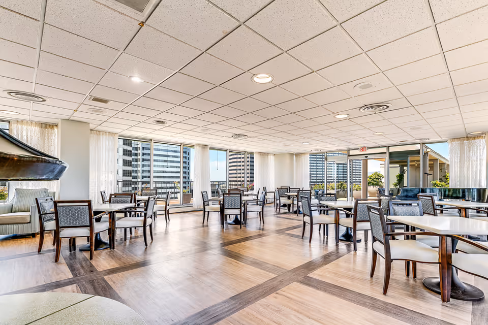 A spacious dining room with multiple tables and chairs arranged neatly. Large windows and glass doors allow natural light to fill the room, offering a view of nearby buildings and an outdoor patio area. The room features a light-colored floor with a darker wood pattern, a white tiled ceiling with recessed lighting, and sheer white curtains on the windows.