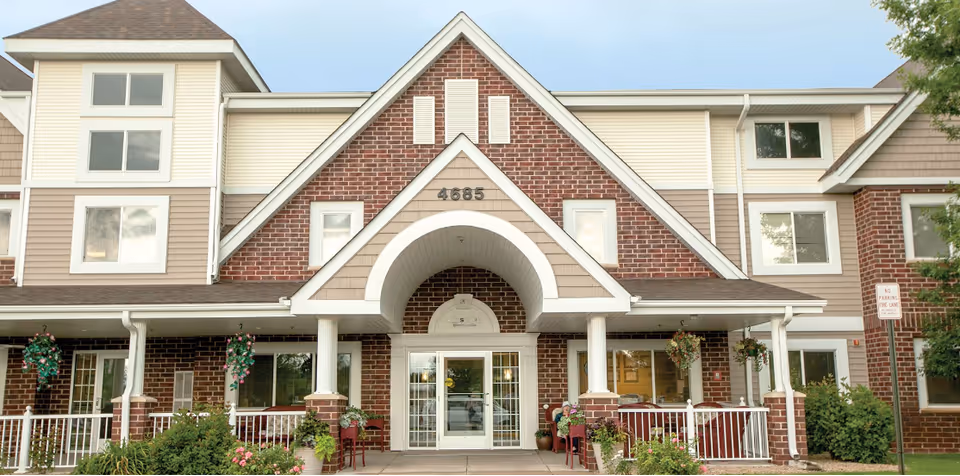 Front exterior view of a senior living facility building with brick and beige siding, a peaked roof entrance with the number 4685 above the door, white columns, hanging flower baskets, and a 'No Parking Fire Lane' sign on the right side.
