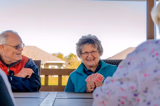 Three elderly people sitting around a table outdoors playing cards, with one woman smiling and holding a hand of cards. The background shows a wooden railing and houses under a clear sky.