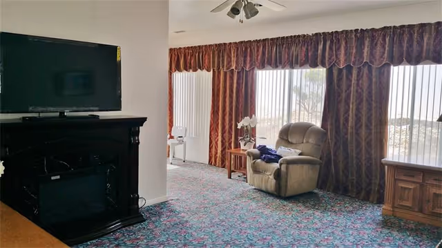 A living room with a wall-mounted TV above a dark mantel, a recliner by large draped windows, patterned carpet, and a ceiling fan.