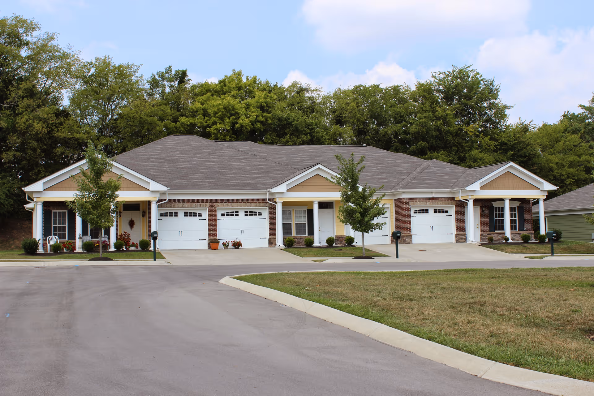 A single-story senior living facility building with multiple attached units, each featuring a garage with white doors, small front porches with columns, and a few small trees and shrubs in front. The building is surrounded by a paved road and grassy areas, with a backdrop of tall green trees under a partly cloudy sky.