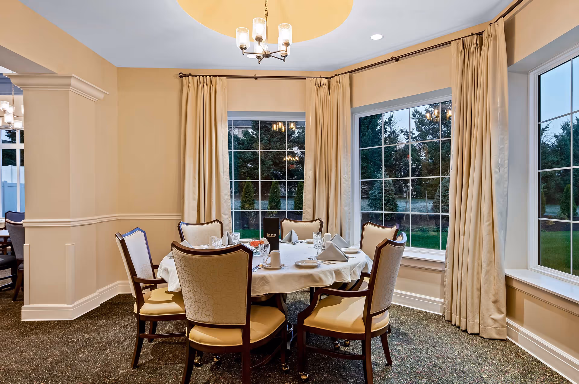 Sunlit dining room with a round table set for six in front of large bay windows and beige curtains.