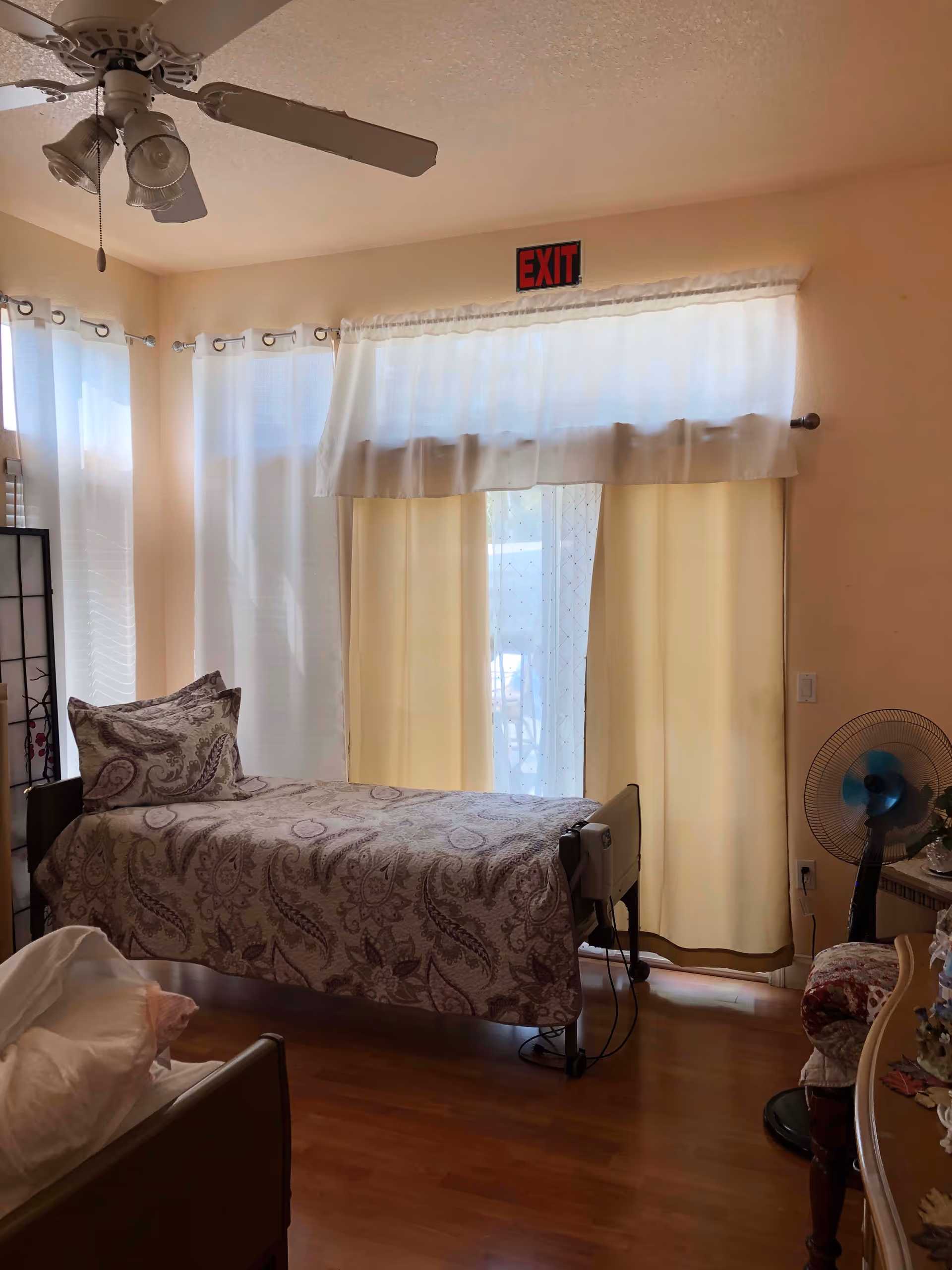 A small bedroom with a single bed covered in a patterned bedspread and a matching pillow. The room has light-colored walls and wooden flooring. There are large windows with sheer white and beige curtains, and an exit sign above the window. A ceiling fan with lights is mounted on the ceiling. A small fan and a chair with a floral cushion are visible near a dresser on the right side of the room.