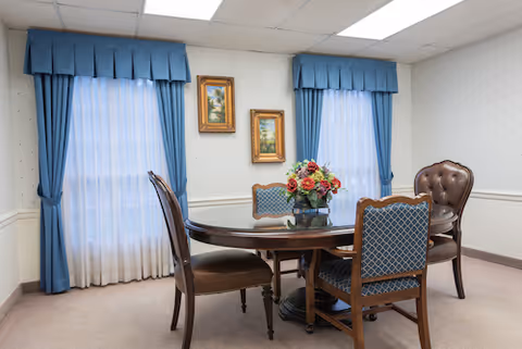 A small dining area with a round wooden table surrounded by four chairs, two with blue patterned upholstery and two with brown leather upholstery. The room has two windows with blue curtains and white sheer drapes. Two framed landscape paintings hang on the wall between the windows. The walls are light-colored with a subtle pattern, and the ceiling has white tiles.