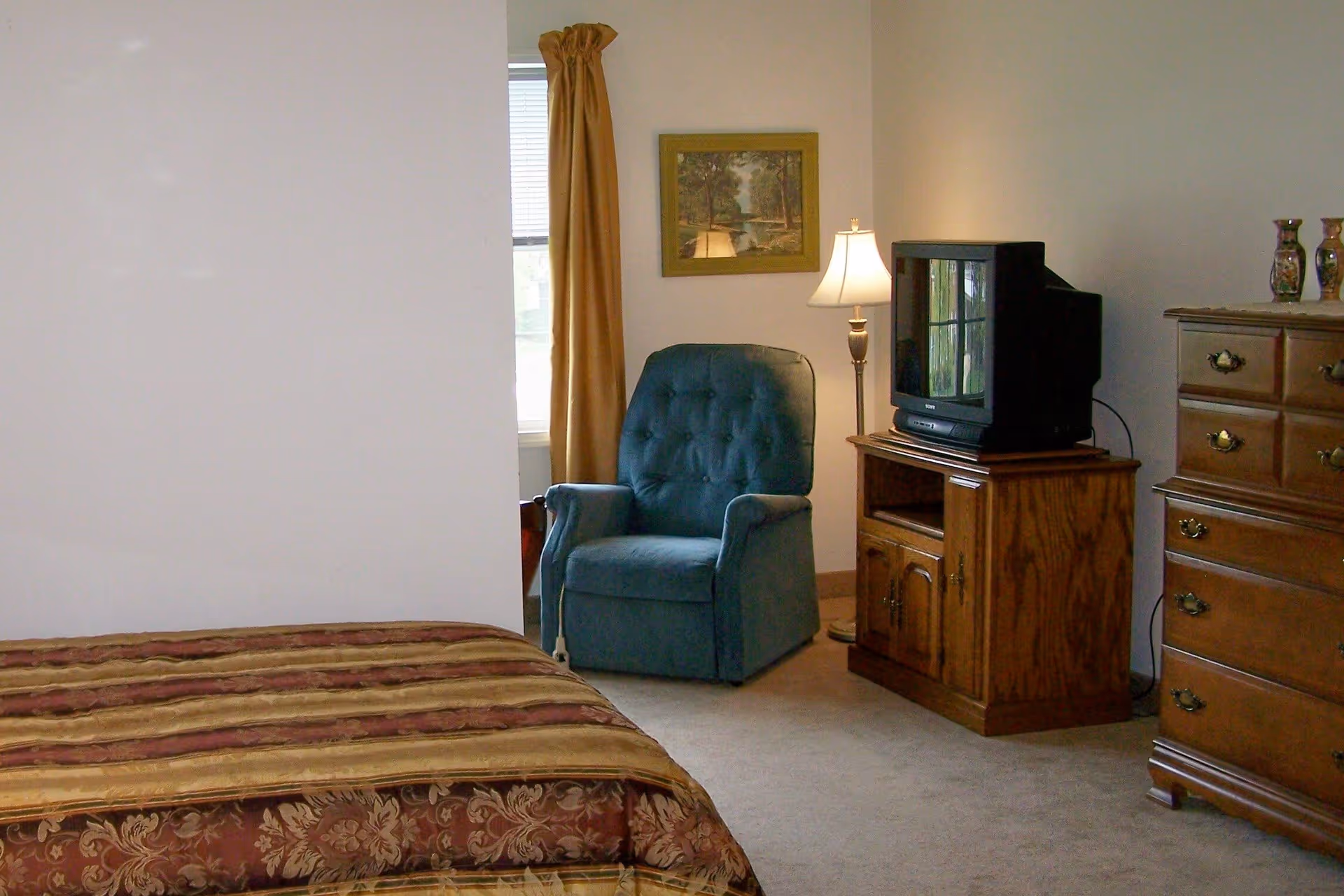 A bedroom with a bed covered in a patterned bedspread, a blue upholstered armchair near a window with gold curtains, a wooden TV stand holding an older model television, a floor lamp, and a wooden dresser with decorative vases on top.