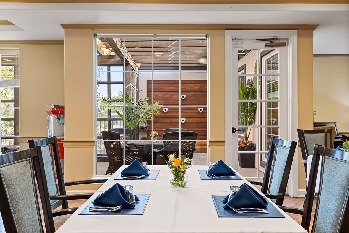 A dining room with a table set for four, featuring blue napkins, glasses, and silverware on blue placemats. The room has large windows and a glass door leading to an outdoor patio area with wicker chairs, plants, and a wooden wall decorated with heart-shaped ornaments.