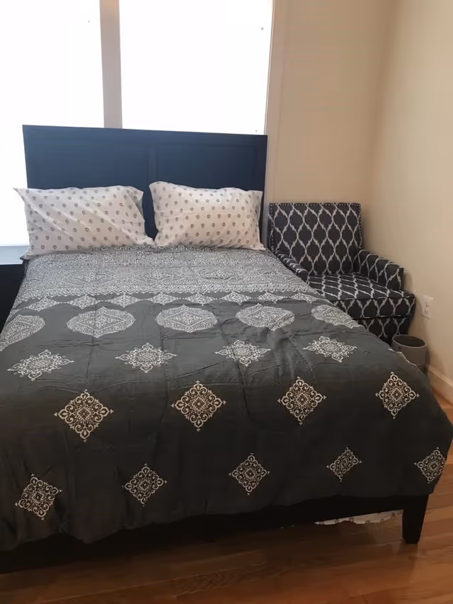 A bedroom with a bed featuring a dark headboard, two white pillows with a small pattern, and a dark bedspread with a white geometric design. To the right of the bed is a patterned armchair with a similar dark and white design. The room has light-colored walls and wooden flooring.