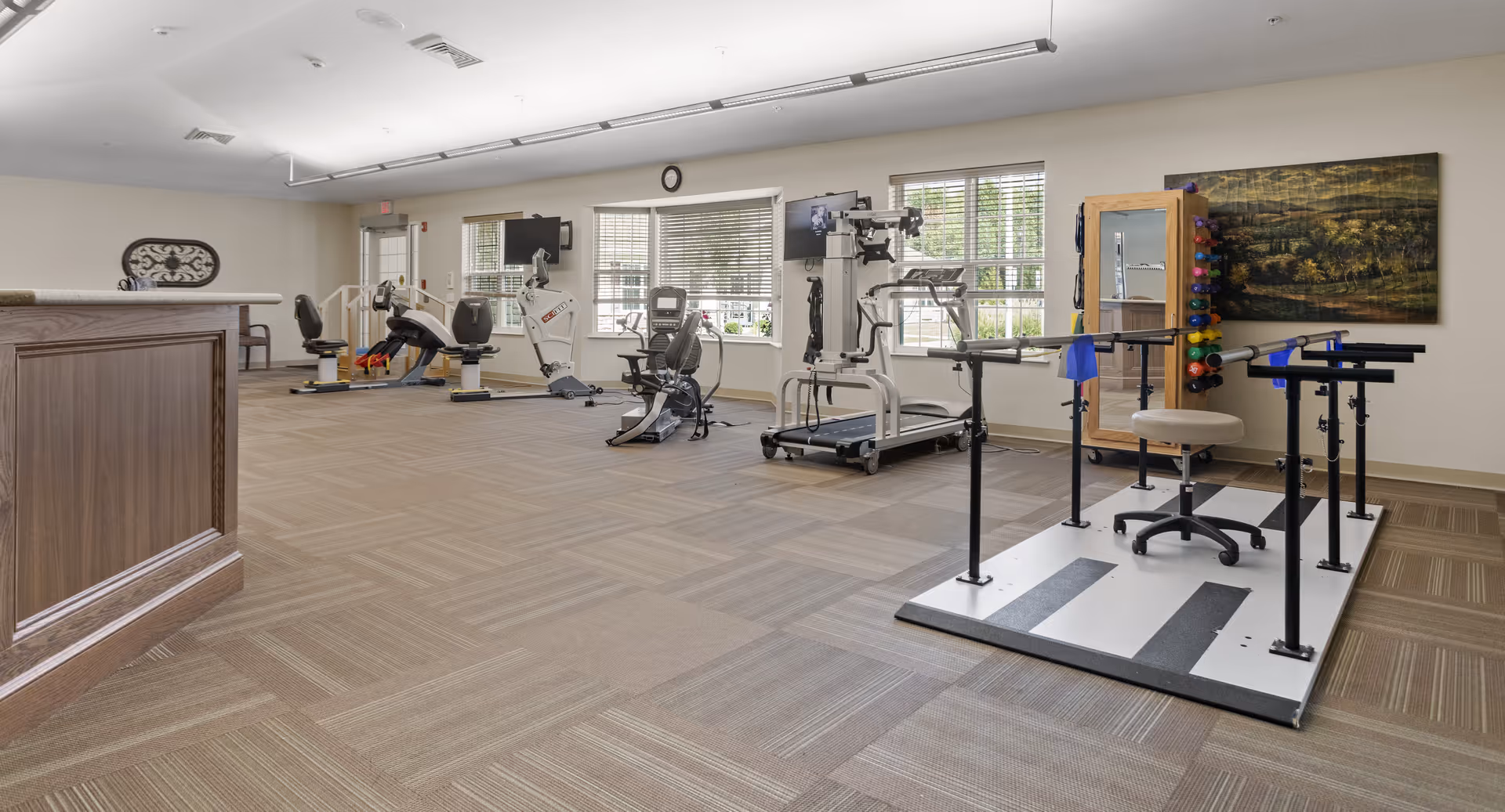 A spacious rehabilitation and exercise room with various fitness equipment including stationary bikes, a treadmill, and parallel bars for physical therapy. The room has large windows allowing natural light, a wooden reception desk on the left, and a painting on the right wall.