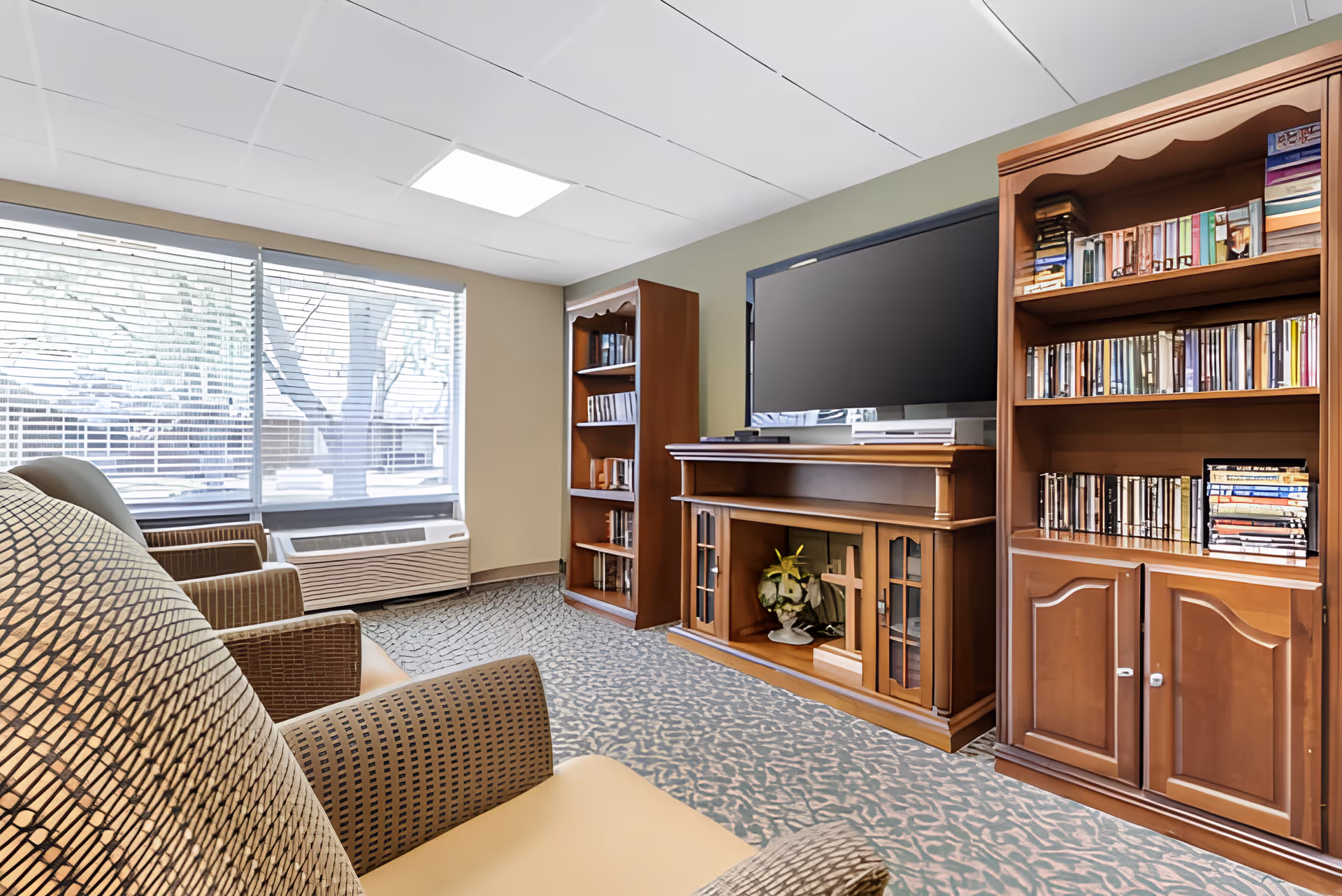 A cozy living room area with patterned armchairs facing a large flat-screen TV on a wooden entertainment center. The entertainment center has glass doors and decorative items inside, including a cross and flowers. On either side of the TV stand are wooden bookshelves filled with books and DVDs. Large windows with blinds allow natural light to fill the room, and the floor is covered with a patterned carpet.